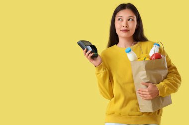 Sarı arka planda kağıt alışveriş torbası ve ödeme terminali olan düşünceli Asyalı genç bir kadın.