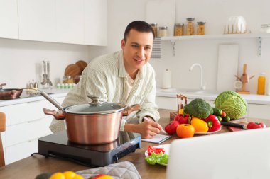 Dizüstü bilgisayarı ve taze sebzeleri olan genç adam mutfakta yemek tarifi yazıyor.