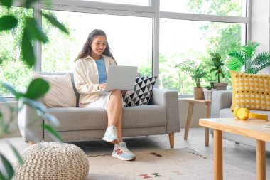 Genç Asyalı kadın evdeki kanepede dizüstü bilgisayar kullanıyor.