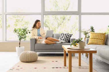 Genç Asyalı kadın evdeki kanepede dizüstü bilgisayar kullanıyor.