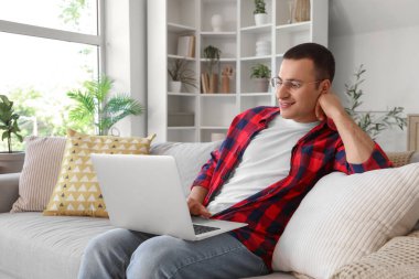 Genç adam evdeki gri koltukta dizüstü bilgisayar kullanıyor.