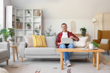 Evdeki gri koltukta oturan dizüstü bilgisayarı olan genç adam.