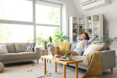 Genç Asyalı kadın evdeki gri kanepede dinleniyor.