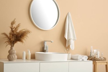 Seramik lavabo, ayna ve vazo. Şık banyoda bej duvarın yanındaki çekmecelerde otlar var.