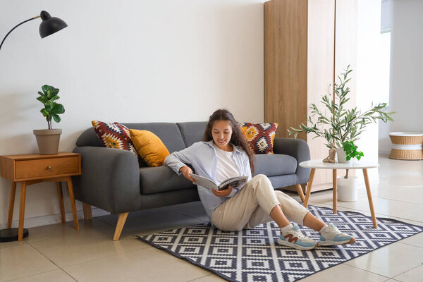 Young Asian woman reading magazine on carpet near black sofa at home
