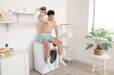 Çamaşır deterjanı şişesiyle evde çamaşır makinesinde oturan genç adam.
