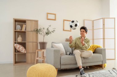 Genç çocuk evdeki kanepede futbol topu atıyor.