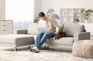 Evdeki kanepede oturan sevimli kedisi ve dizüstü bilgisayarı olan genç adam.