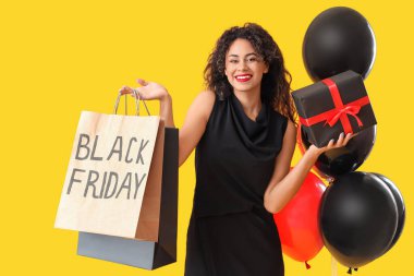 Alışveriş torbaları, hediye kutuları ve sarı arka planda balonları olan güzel bir Afro-Amerikalı kadın. Kara Cuma indirimi