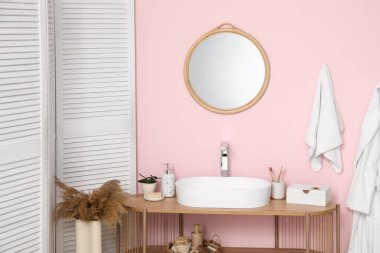 Modern sink with bath supplies and houseplant on shelving unit near pink wall in bathroom