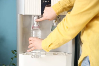Plastik şişeyi mavi duvarın yanındaki soğutucudan filtreli suyla dolduran kadın.
