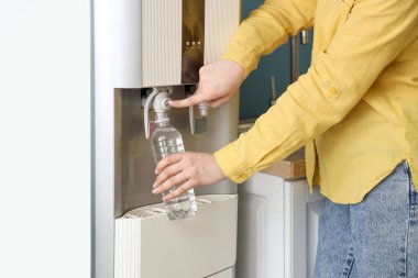 Plastik şişeyi mavi duvarın yanındaki soğutucudan filtreli suyla dolduran kadın.