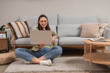 Evdeki gri kanepenin yanında dizüstü bilgisayar kullanan genç bir kadın.