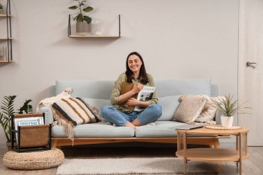 Genç bir kadın, evdeki duvarın yanındaki gri koltukta dergi okuyor.