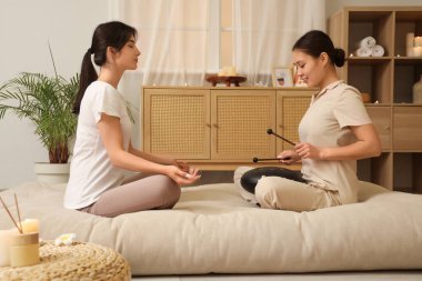 Thai therapist playing glucophone with meditating young woman in spa salon