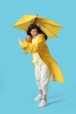 Young woman in raincoat with broken umbrella and strong wind on blue background