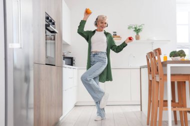 Mutfakta orta yaşlı kulaklıklı ve sebzeli bir kadın dans ediyor.