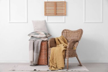 Beyaz duvarın yanında yastık yığını olan hasır koltuk ve sepet.