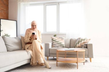 Başörtülü şık Müslüman kadın evdeki koltukta cep telefonu kullanıyor.