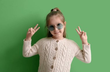 Güneş gözlüklü ve kazaklı şirin kız yeşil arka planda zafer jesti gösteriyor.