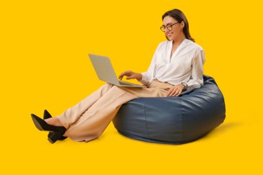 Sarı arka planda fasulye koltukta otururken dizüstü bilgisayarla çalışan güzel bir genç kadın.