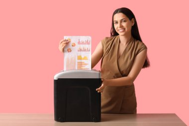 Pembe arka planda kağıt öğütücüyle belgeleri yok eden mutlu genç kadın.