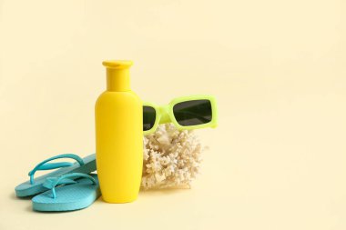 Bir şişe güneş kremi, parmak arası terlik, bej arka planda mercan ve güneş gözlüğü.