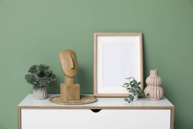 Yeşil duvarın yanında biblolar, ev bitkileri ve fotoğraf çerçeveleri olan modern bir giysi.