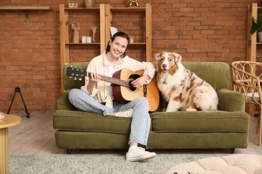 Güzel genç bir bayan evde hoş bir Avustralya çoban köpeğiyle gitar çalıyor.