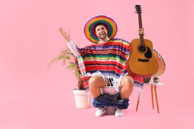 Gitarlı Meksikalı genç adam pembe arka planda klozetin üzerinde oturuyor.