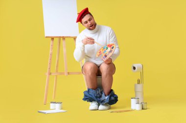 Boya paleti ve fırçası olan erkek sanatçı sarı arka planda klozetin üzerinde oturuyor.