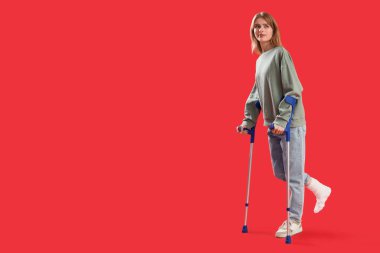 Kırık ayak ve kırmızı arka planda koltuk değnekleriyle kaza sonucu yaralanan genç bir kadın.