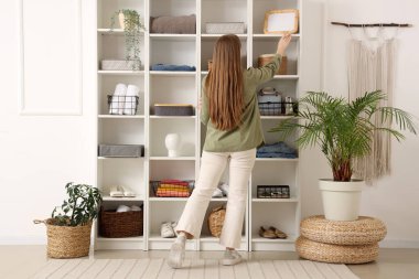 Rafların yanında genç bir kadın var. Giysileri ve dekorları evde.