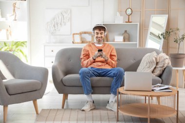 Kulaklıklı, cep telefonlu, evdeki kanepede müzik dinleyen genç dövmeli adam.