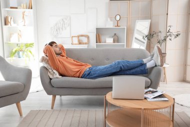 Kulaklıklı genç adam evdeki kanepede müzik dinliyor.