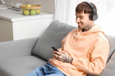 Kulaklıklı ve cep telefonlu genç dövmeli adam mutfaktaki kanepede müzik dinliyor.