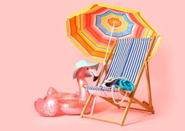 Renkli şemsiyeli çizgili iskemle, şişirilebilir yüzük ve pembe arka planda plaj aksesuarları.
