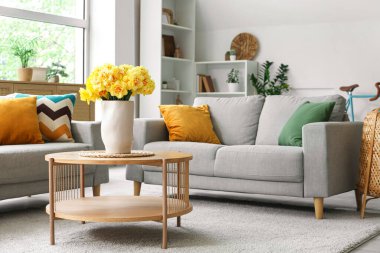 Aydınlık oturma odasının içinde gri kanepeler ve ahşap sehpanın üzerinde sarı narsisli çiçekler.