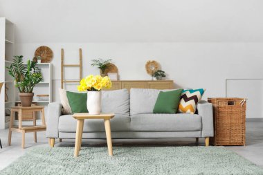 Oturma odasının içinde gri kanepe ve ahşap sehpada sarı narsisli çiçekler.