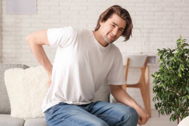 Handsome young man sitting on sofa and suffering from back pain at home