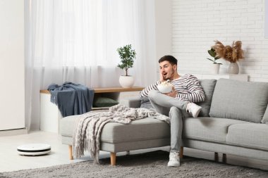 Young man watching movie with modern robot vacuum cleaner at home