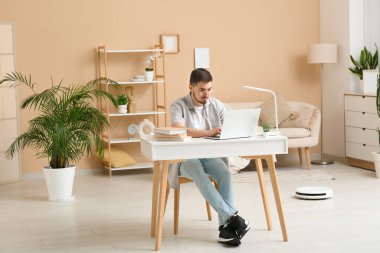 Young man using laptop at table with modern robot vacuum cleaner on floor in home office