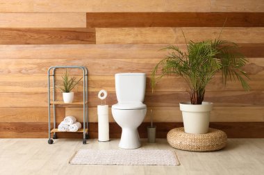 Interior of restroom with toilet bowl, shelving unit and houseplant near wooden wall clipart