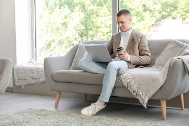 Modern ev ofisindeki kanepede oturan dizüstü bilgisayarı ve bir fincan kahvesiyle serbest çalışan genç bir erkek.