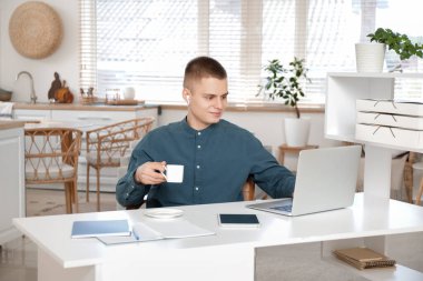 Hafif ev ofisinde dizüstü bilgisayar kullanan bir fincan kahveli serbest çalışan genç bir erkek.