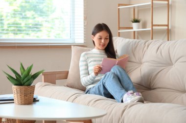 Evdeki kanepede kitap okuyan güzel genç bir kadın.