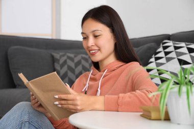 Evdeki kanepenin yanında kitap okuyan güzel genç bir kadın.