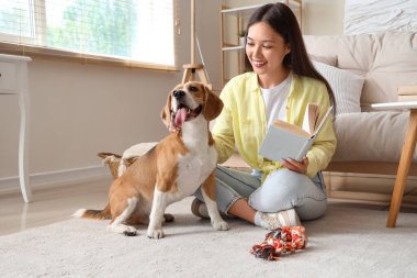 Genç bir kadın elinde kitapla oturma odasında yerde tatlı bir Beagle köpeğiyle oynuyor.