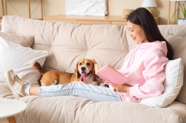 Genç bir kadın kitap okuyor ve oturma odasındaki kanepede sevimli Beagle köpeğini okşuyor.