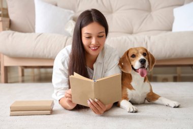 Genç bir kadın yerde uzanıyor ve oturma odasında tapılası Beagle köpeğiyle kitap okuyor.